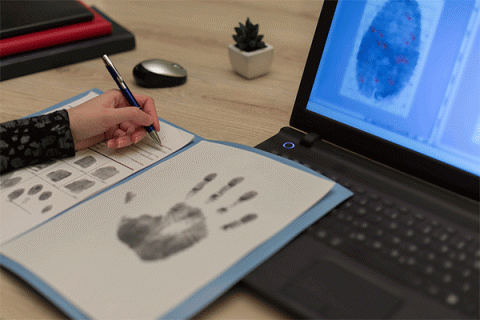 Detailed image of the hand of a student writing on a form containing a full handprint and fingerprints while viewing a fingerprint on a laptop.